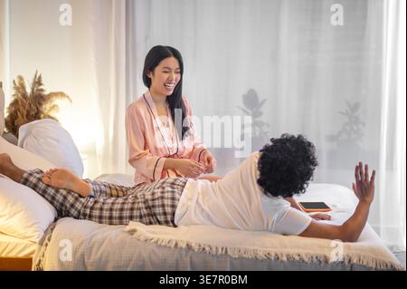 Donna ispanica sorridente che ride seduta vicino al fidanzato sdraiato sul letto di casa Foto Stock