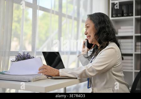 Donna d'affari asiatica che riceve una telefonata che controlla i documenti che guardano lontano mentre si siede a un tavolo con un portatile in ufficio Foto Stock