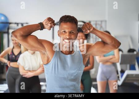 Un uomo nero sorridente e muscoloso che flessa i muscoli del braccio mentre si trova di fronte ad amici femminili in palestra Foto Stock