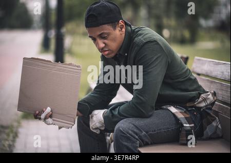 Un senzatetto nero con un cappello che tiene un cartone vuoto che guarda in basso mentre si siede sulla panchina per strada durante il giorno Foto Stock