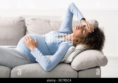 Stress donna incinta di razza mista con mal di testa sdraiata sul divano di casa Foto Stock