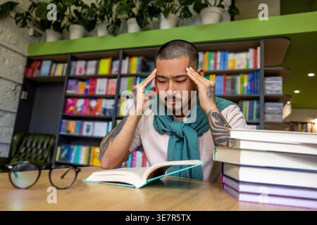 Uomo asiatico tatuato con gli occhi chiusi che toccano i suoi templi seduto a tavola con libri in biblioteca Foto Stock