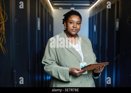 Ingegnere IT donna seria e nera che tiene il tablet in piedi nella sala server del data center Foto Stock