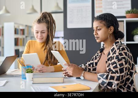 Studentesse di razza mista e caucasiche che studiano usando tablet seduti a tavola insieme in biblioteca Foto Stock