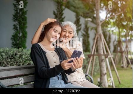 Donna thailandese che si diverte a condividere lo smartphone con la nonna mentre si siede su una panchina di legno nel parco durante il giorno Foto Stock