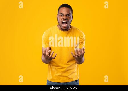 Uomo nero che urla a mani aperte mentre si trova su sfondo giallo in studio Foto Stock