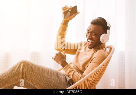 Uomo nero che ride indossando le cuffie che tengono lo smartphone seduto sulla sedia vicino alla finestra di casa Foto Stock