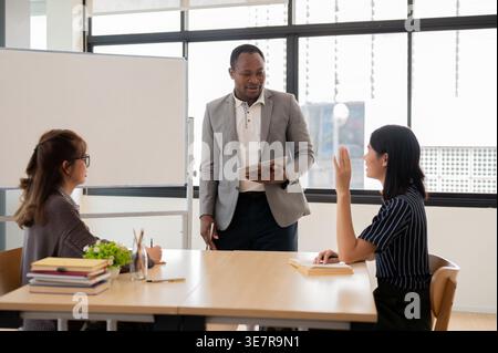 Uomo d'affari nero sorridente che tiene in mano un tablet mentre è in piedi e conduce la riunione del team con colleghi asiatici in ufficio Foto Stock