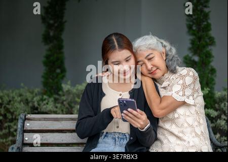 Donna thailandese che si diverte a condividere lo smartphone con la nonna mentre si siede su una panchina di legno nel parco durante il giorno Foto Stock