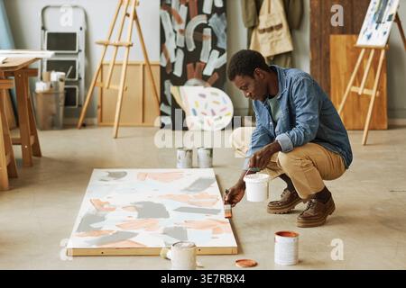 Artista maschile nero che dipinge su tela mentre accovacciato sul pavimento in studio d'arte Foto Stock