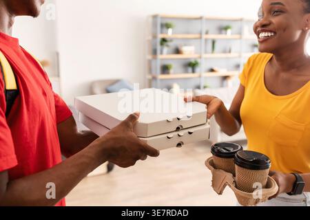 Donna nera sorridente che riceve scatole per pizza e tazze da caffè usa e getta dal fattorino mentre si trova davanti alla porta di casa Foto Stock
