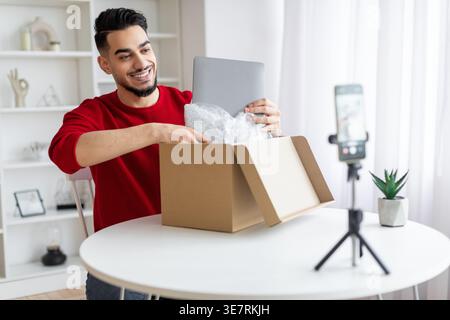 Influencer maschile arabo che registra contenuti video utilizzando lo smartphone che disimballano il pacco con il laptop seduto al tavolo di casa Foto Stock