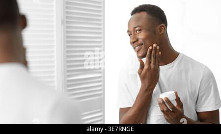 Uomo nero in camicia bianca che regge il vaso idratante e applica la crema sul viso stando in piedi davanti allo specchio nel bagno di casa Foto Stock