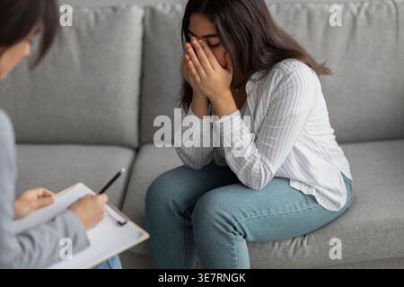 Donna mediorientale che piange mentre siede sul divano di fronte a una psicologa che prende appunti in clinica Foto Stock