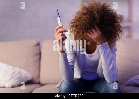 Donna nera depressa con capelli ricci che toccano la testa tenendo il kit per il test di gravidanza seduto sul divano nel soggiorno di casa Foto Stock