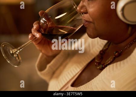 Donna nera di mezza età che ascolta la musica con le cuffie e beve vino mentre è in salotto a casa Foto Stock