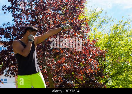Uomo ispanico con canotta nera e pantaloncini verdi che praticano tecniche di pugilato nel parco nelle giornate di sole Foto Stock