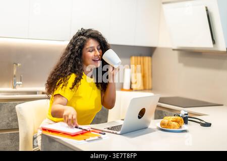 Donna ispanica con capelli ricci che partecipa a una riunione online utilizzando un computer portatile mentre fa colazione al bancone della cucina a casa Foto Stock