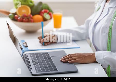Immagine ritagliata di una nutrizionista nera che lavora su un laptop e scrive la storia dei clienti mentre si siede a tavola con una ciotola di frutta e verdura io... Foto Stock