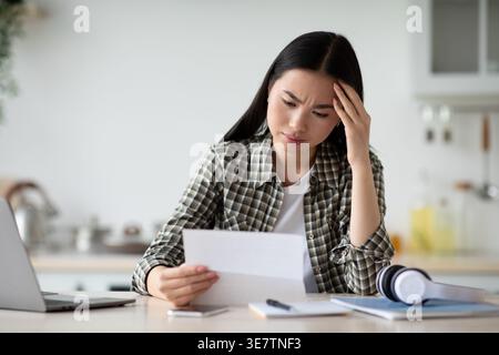 Donna asiatica che legge una lettera e tocca la fronte mentre siede a un tavolo con le cuffie e il portatile a casa Foto Stock