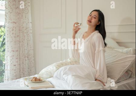 Sorridente donna asiatica nella camicia da notte che spruzza profumo sul collo seduta a letto con colazione in camera da letto a casa Foto Stock