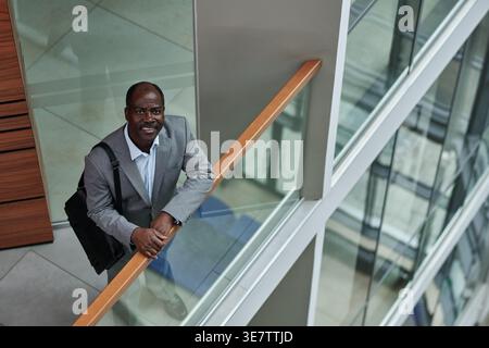 Uomo d'affari nero di mezza età con borsa a spalla in piedi e braccia appoggiate sulla ringhiera nel balcone dell'ufficio Foto Stock