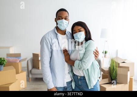 Coppia nera che indossa maschere protettive per il viso che si abbraccia mentre sta in piedi tra le scatole di cartone nel soggiorno di casa Foto Stock