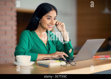 Donna indiana che indossa una cuffia per videochiamare utilizzando un computer portatile seduto al tavolo del bar Foto Stock