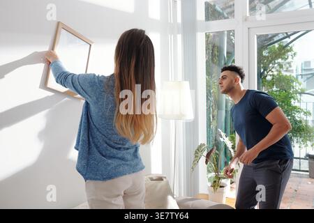 Uomo di razza mista che guarda la cornice impiccata dalla ragazza caucasica nel soggiorno di casa Foto Stock