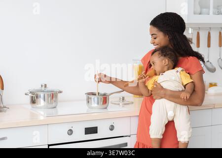 Madre nera che cucina il cibo trasportando il bambino mentre si trova in cucina a casa Foto Stock