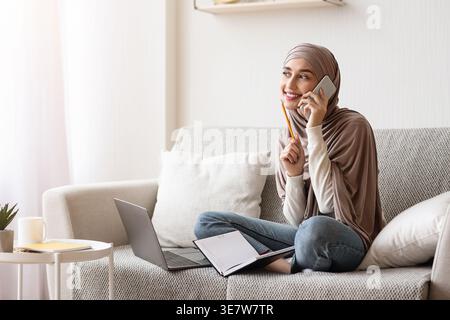 Donna musulmana araba in hijab che ha una telefonata con una penna seduta con le gambe incrociate sul divano con un portatile e un notebook a casa Foto Stock
