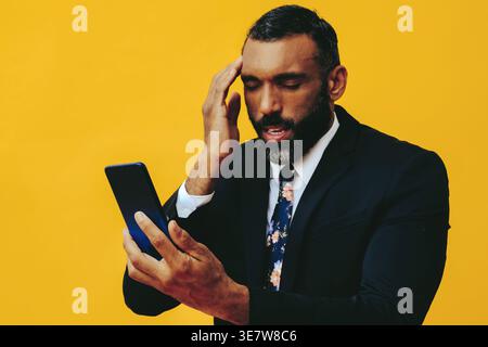 Uomo d'affari barbuto e stressato in costume e cravatta, videochiamata sullo smartphone e toccando il tempio in piedi su sfondo giallo in... Foto Stock