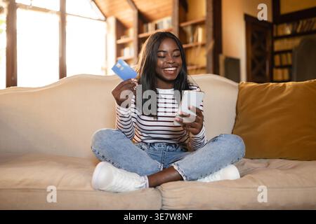 Adolescente nera che tiene la carta di credito mentre utilizza lo smartphone seduto con le gambe incrociate sul divano nel soggiorno di casa Foto Stock