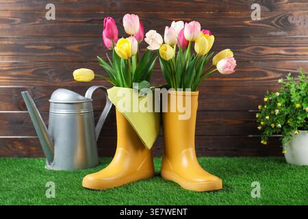 Splendidi fiori di tulipani in stivali di gomma gialli e lattine da innaffiare sull'erba artificiale vicino alle pareti di legno Foto Stock