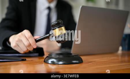 Avvocato uomo asiatico che tiene in mano il martelletto del giudice mentre si siede al tavolo con il portatile in ufficio Foto Stock