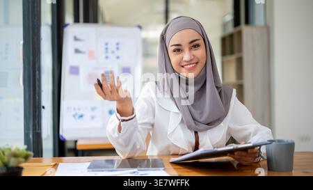 Una donna d'affari musulmana asiatica sorridente che indossa un hijab siede alla sua scrivania in un ufficio moderno. Foto Stock