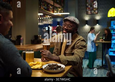 Uomo nero con occhiali che reggono un bicchiere di birra guardando gli amici seduti al tavolo del bar Foto Stock