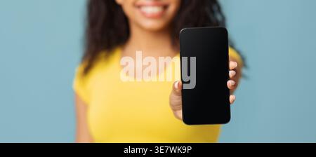Donna nera sorridente che indossa una camicia gialla che mostra lo smartphone mentre è in piedi su sfondo blu in studio Foto Stock