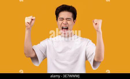Uomo asiatico che indossa una camicia bianca urlando e stringendo i pugni mentre sta in piedi con gli occhi chiusi contro il muro arancione in studio Foto Stock