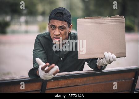 Un senzatetto nero con un cappello che tiene in mano un cartone vuoto mentre sta in piedi a chiedere soldi per strada durante il giorno Foto Stock