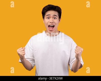 Uomo asiatico che indossa una camicia bianca urlando e stringendo i pugni mentre sta in piedi contro il muro arancione in studio Foto Stock