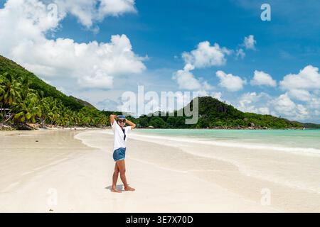 Un viaggiatore sorge sulla splendida spiaggia di sabbia bianca di Anse Source d'Argent nelle Seychelles, crogiolandosi al sole. Le onde serene e la vegetazione lussureggiante creano lo sfondo perfetto per le vacanze di relax. Foto Stock