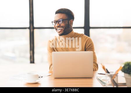Allegro uomo nero che indossa occhiali da vista che guarda lontano seduto a un tavolo con un computer portatile in ufficio Foto Stock