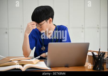 Studente di medicina maschile asiatico stressato in scrub blu toccando la fronte guardando in basso mentre si siede a tavolo in spogliatoio Foto Stock