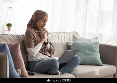 Una donna araba scioccata con le mani schiacciate che indossano un hijab che guarda lo schermo del notebook mentre è seduta con le gambe incrociate sul divano nel soggiorno di casa Foto Stock