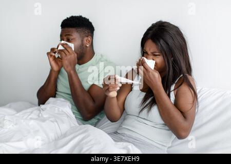 Coppia nera con febbre che misura la temperatura corporea con il termometro che soffia il naso usando il tovagliolo seduto sul letto di casa Foto Stock