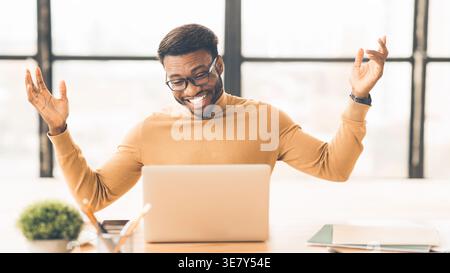 Allegro uomo nero che indossa occhiali da vista guardando un computer portatile seduto a un tavolo a braccia aperte in ufficio Foto Stock