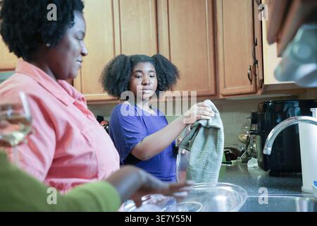 La figlia nera con afro che usa un asciugamano da cucina nelle piastre di asciugatura aiutando sua madre mentre si trova vicino al lavandino della cucina a casa Foto Stock