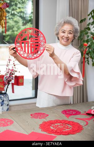 Sorridente donna cinese anziana in piedi a tavola con carta rossa per il Capodanno cinese tagliata a casa Foto Stock