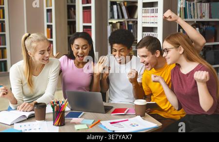 Allegro e variegato gruppo di studenti universitari che festeggiano davanti a un notebook in biblioteca Foto Stock